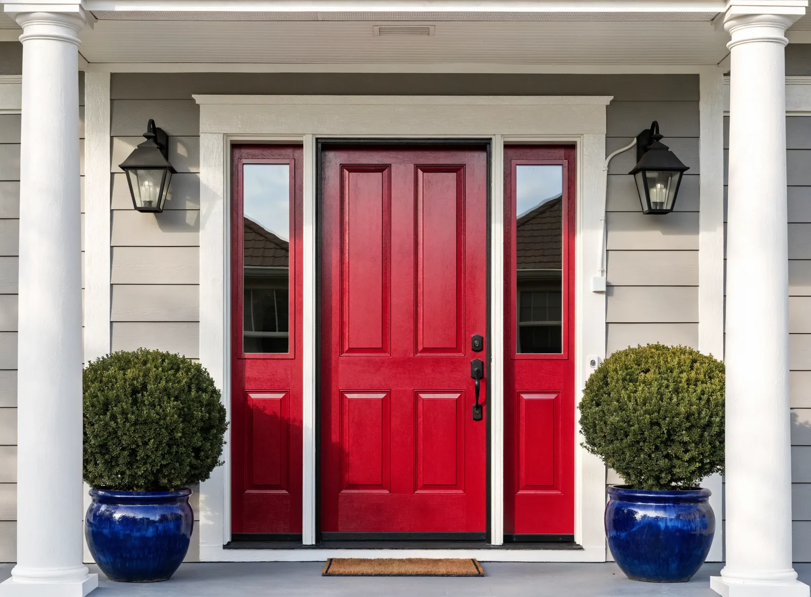 An upscale front entry: a glossy red door with sidelight windows, black lanterns, boxwoods in blue pots, and a doormat